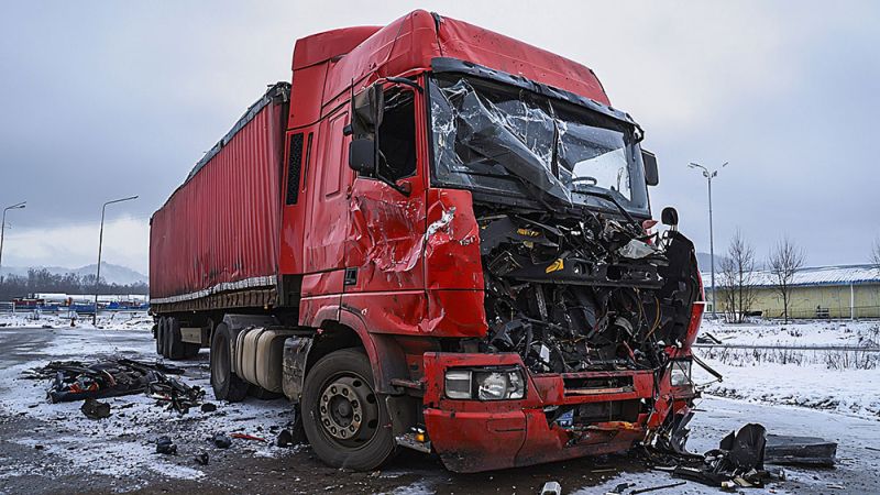 Crashed red truck in snowy area, showing significant front-end damage and debris around, overcast sky in the background.