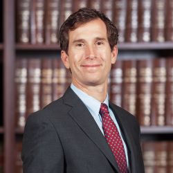 Man in a suit and red tie smiling in front of bookshelves filled with brown books.