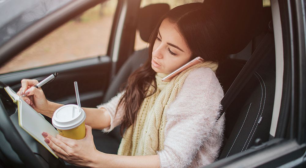 Young woman multitasking in car, talking on phone, writing notes, holding coffee cup, showcasing busy lifestyle.