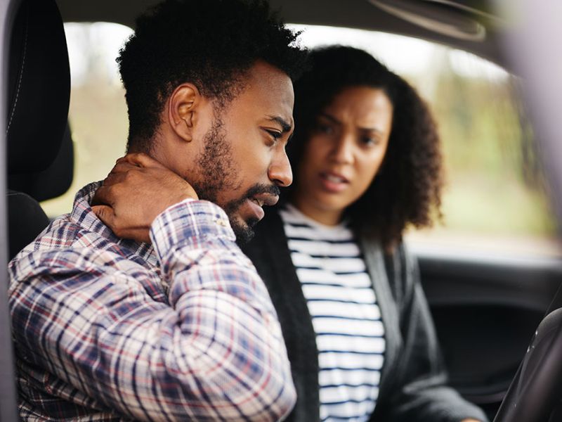 Man in a car holding neck in pain while concerned woman looks on, highlighting accident aftermath.
