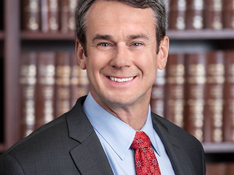 Smiling man in a suit and red tie in front of a bookshelf filled with brown books.