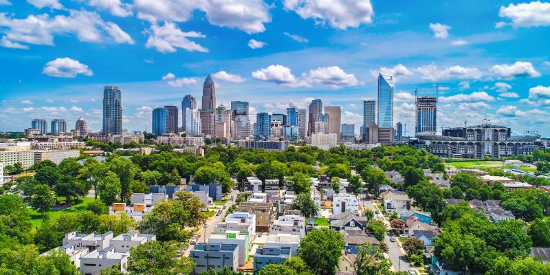 Skyline of a vibrant cityscape with tall buildings, lush greenery, and scattered clouds under a bright blue sky.