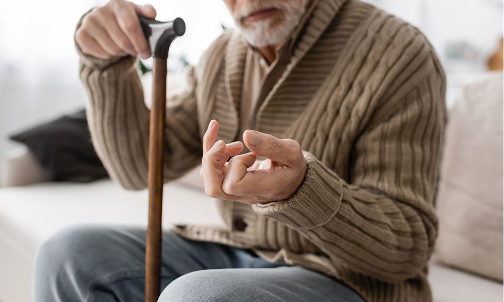 Elderly man with cane experiencing hand pain while seated, wearing a brown sweater, highlighting arthritis issues.