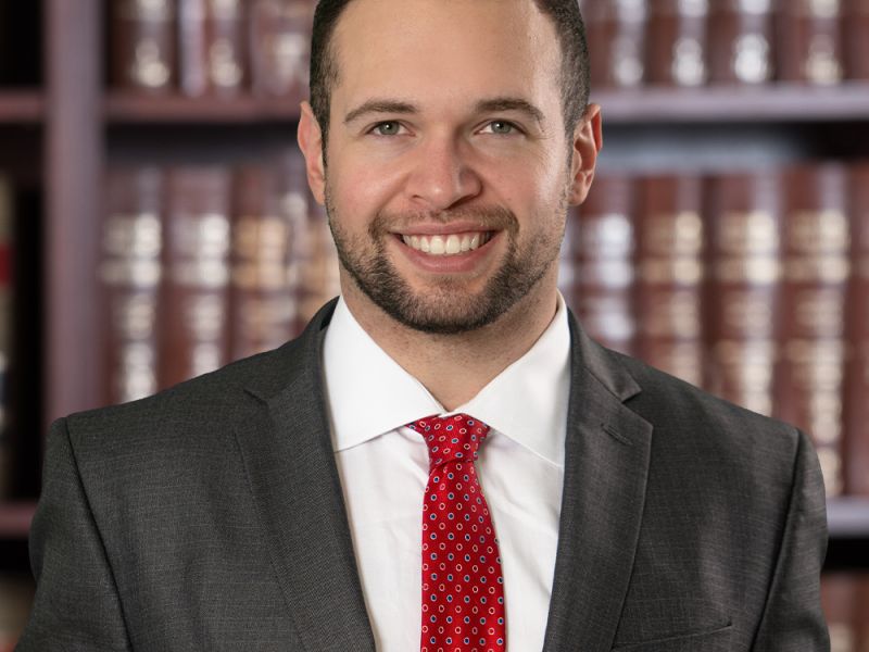 Smiling man in a suit and red tie stands before a bookshelf with law books, conveying professionalism and confidence.