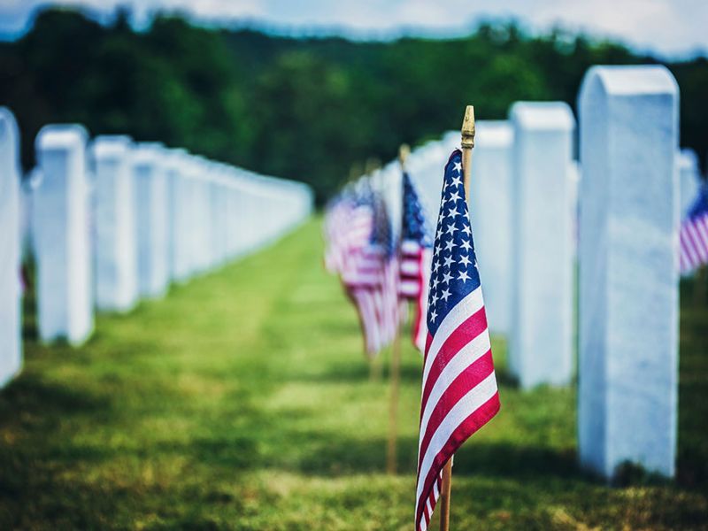 American flags adorn rows of white headstones in a solemn cemetery setting under a blue sky.