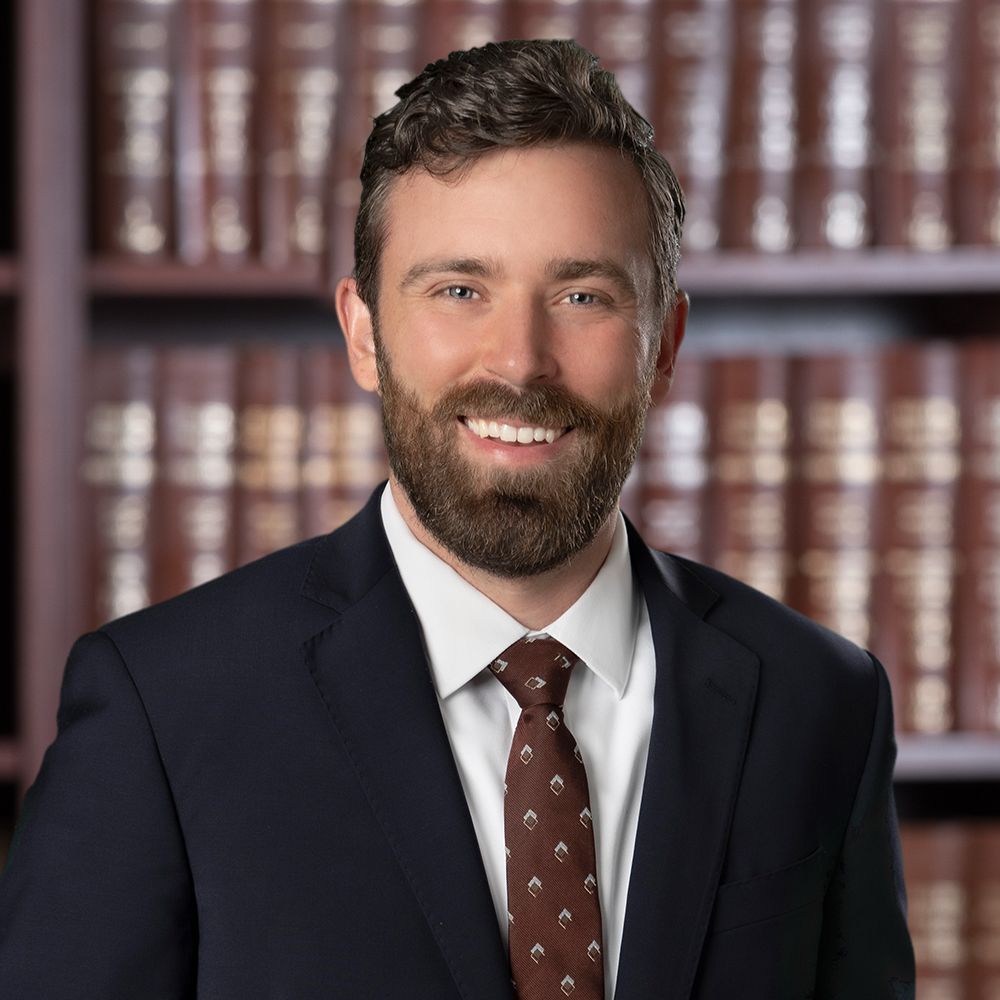 A smiling man in a suit with a beard stands in front of a bookshelf with books in a professional setting.