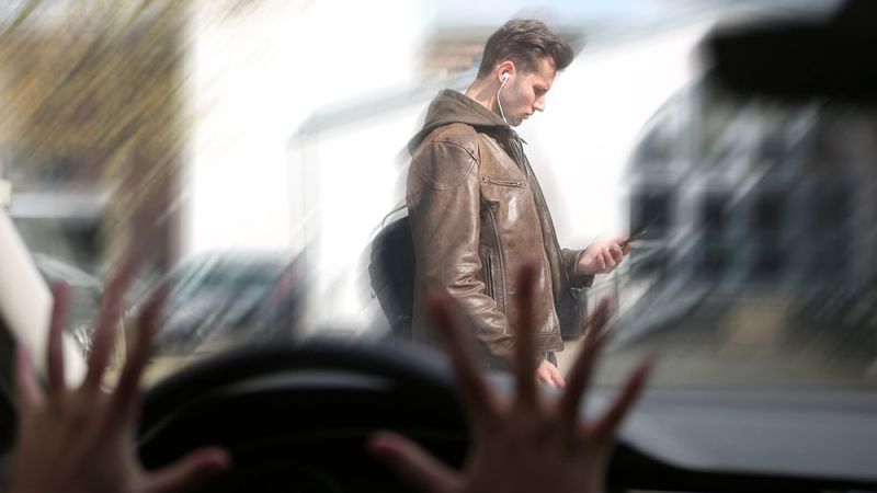 Pedestrian using phone while crossing street, viewed from car driver's perspective, highlighting road safety issues.