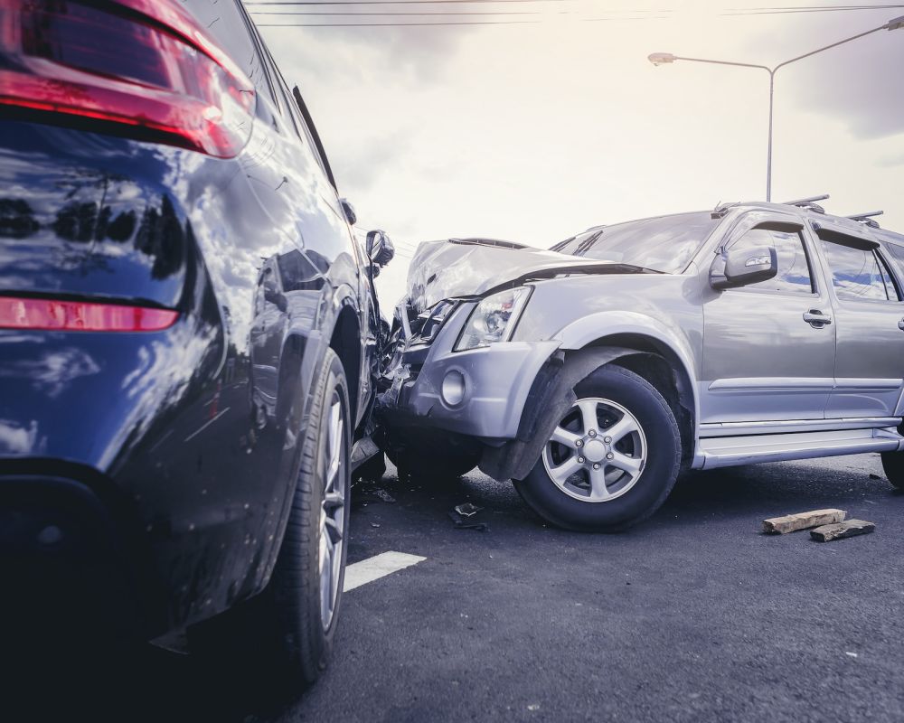 Two SUV cars in a collision on a road, showing damaged front ends under a bright sky.