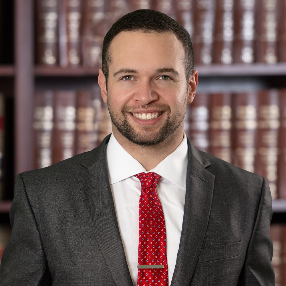 Smiling man in a suit and red tie stands before a bookshelf with law books, conveying professionalism and confidence.