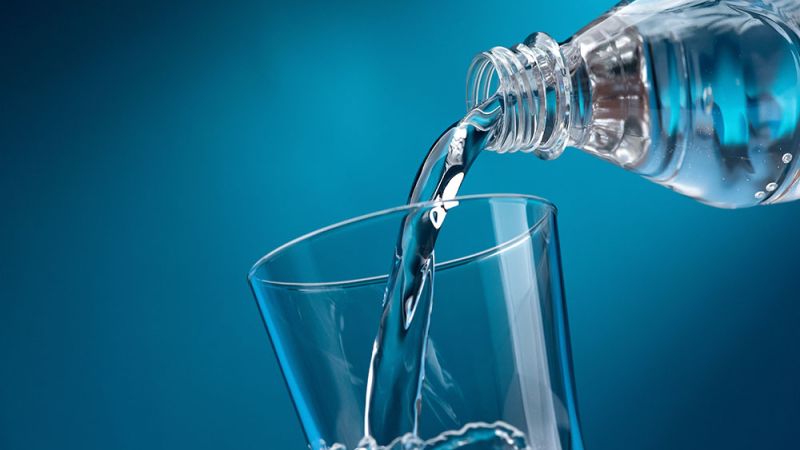 Pouring clear water from a bottle into a glass against a blue background, symbolizing hydration and purity.