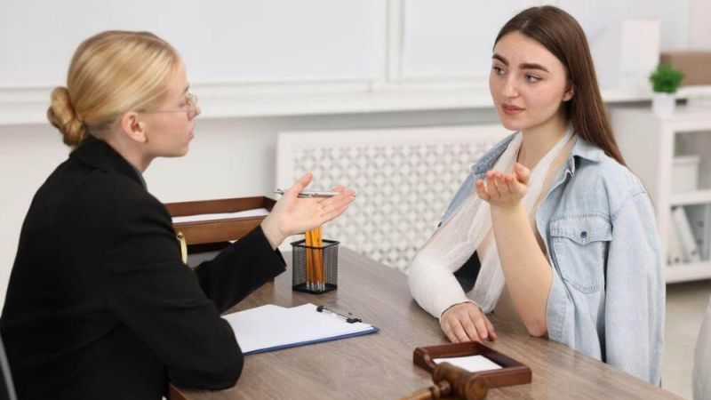 Lawyer counseling a young woman with a cast on her arm at a desk, gavel and documents visible on the table, in a legal office.