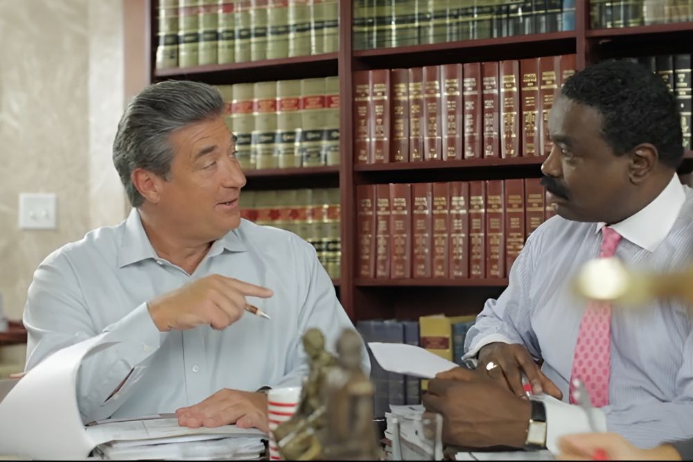 Two men discussing documents in an office with law books on a shelf in the background.