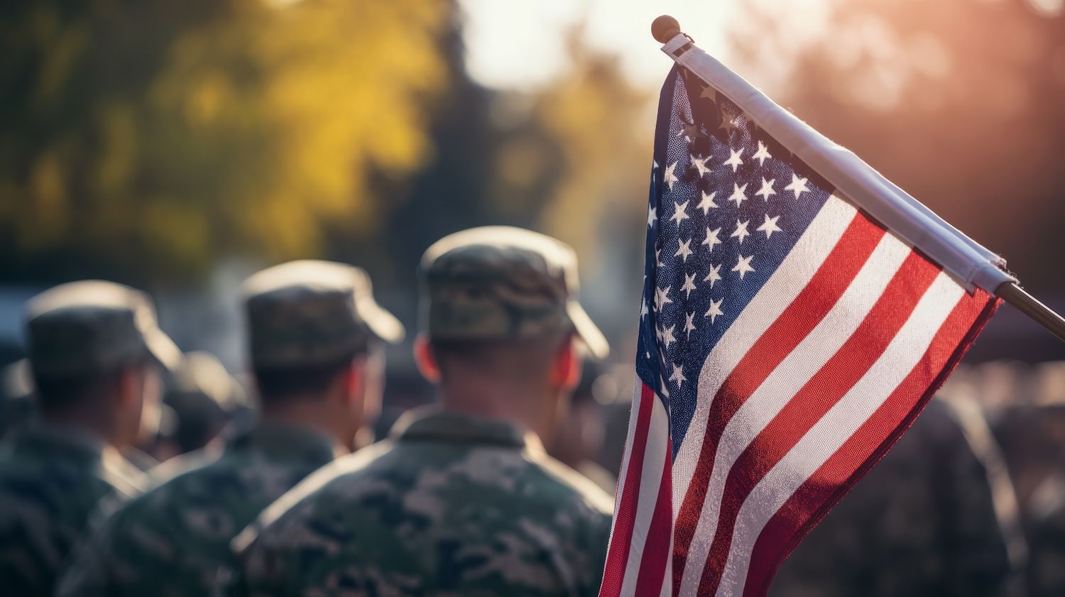 American flag waving in front of soldiers in uniform, with sunlight filtering through trees.