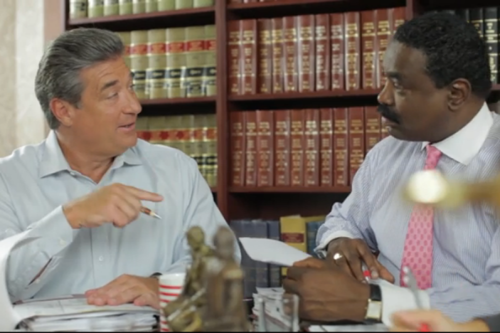 Two businessmen discussing work at a desk filled with papers and books in a law office setting.