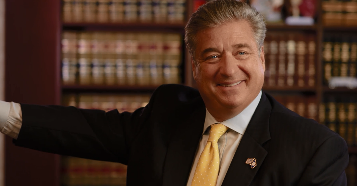 Smiling man in a suit with a yellow tie, standing in front of bookshelves filled with legal books.