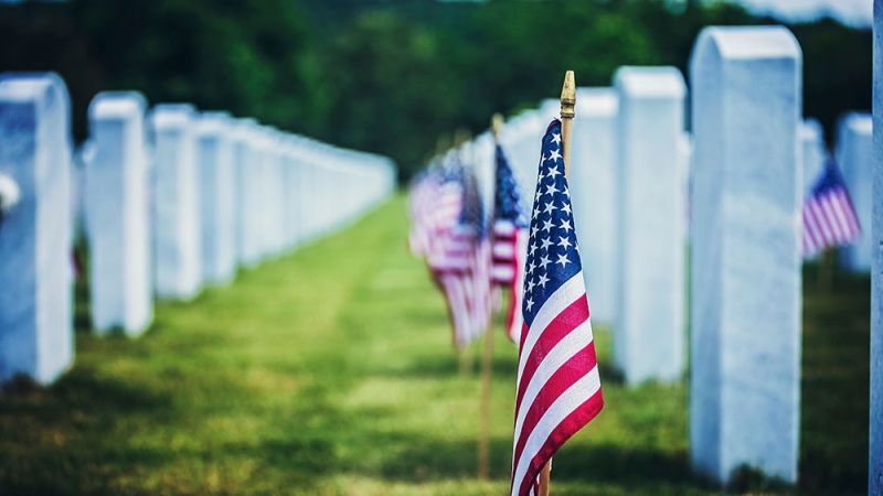 American flags adorn rows of white headstones in a solemn cemetery setting under a blue sky.