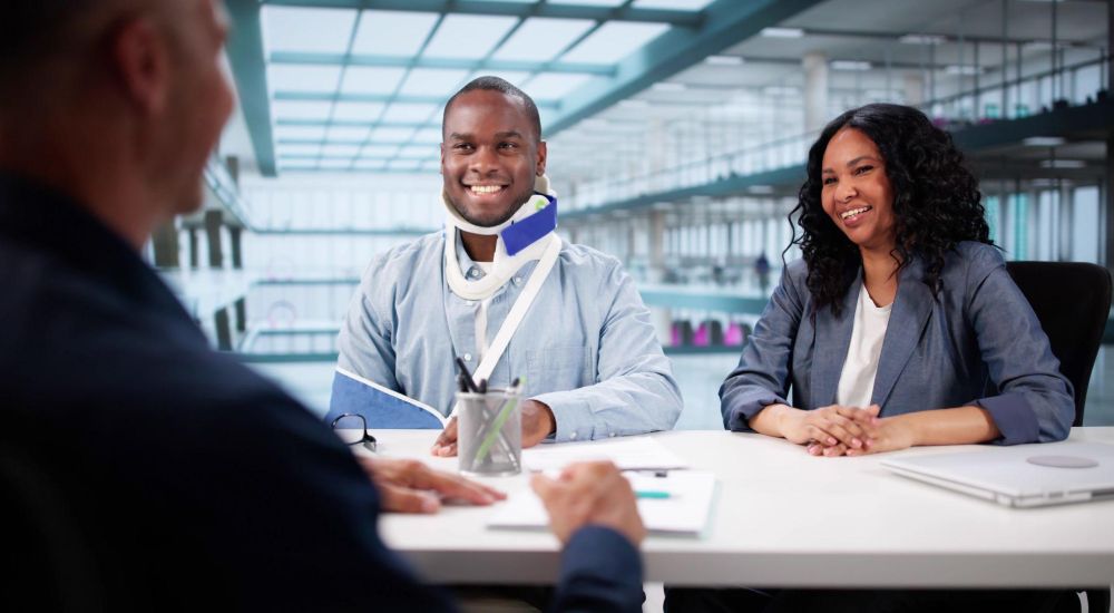 Meeting with a smiling man in a neck brace and woman at a modern office table, discussing documents.