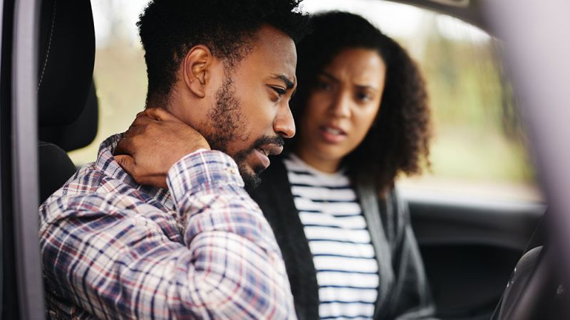 Man in a car holding neck in pain while concerned woman looks on, highlighting accident aftermath.