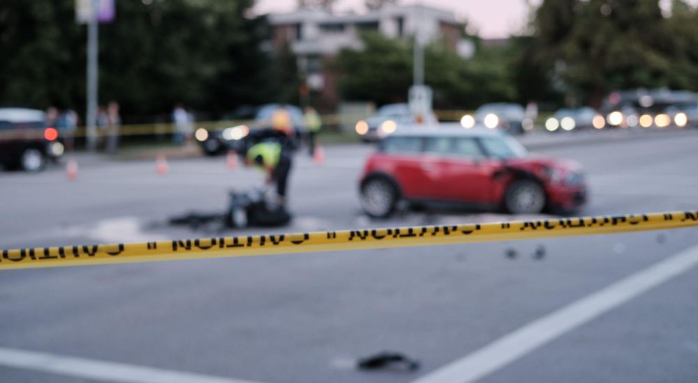 Yellow caution tape at car accident scene with police and damaged red vehicle in background.