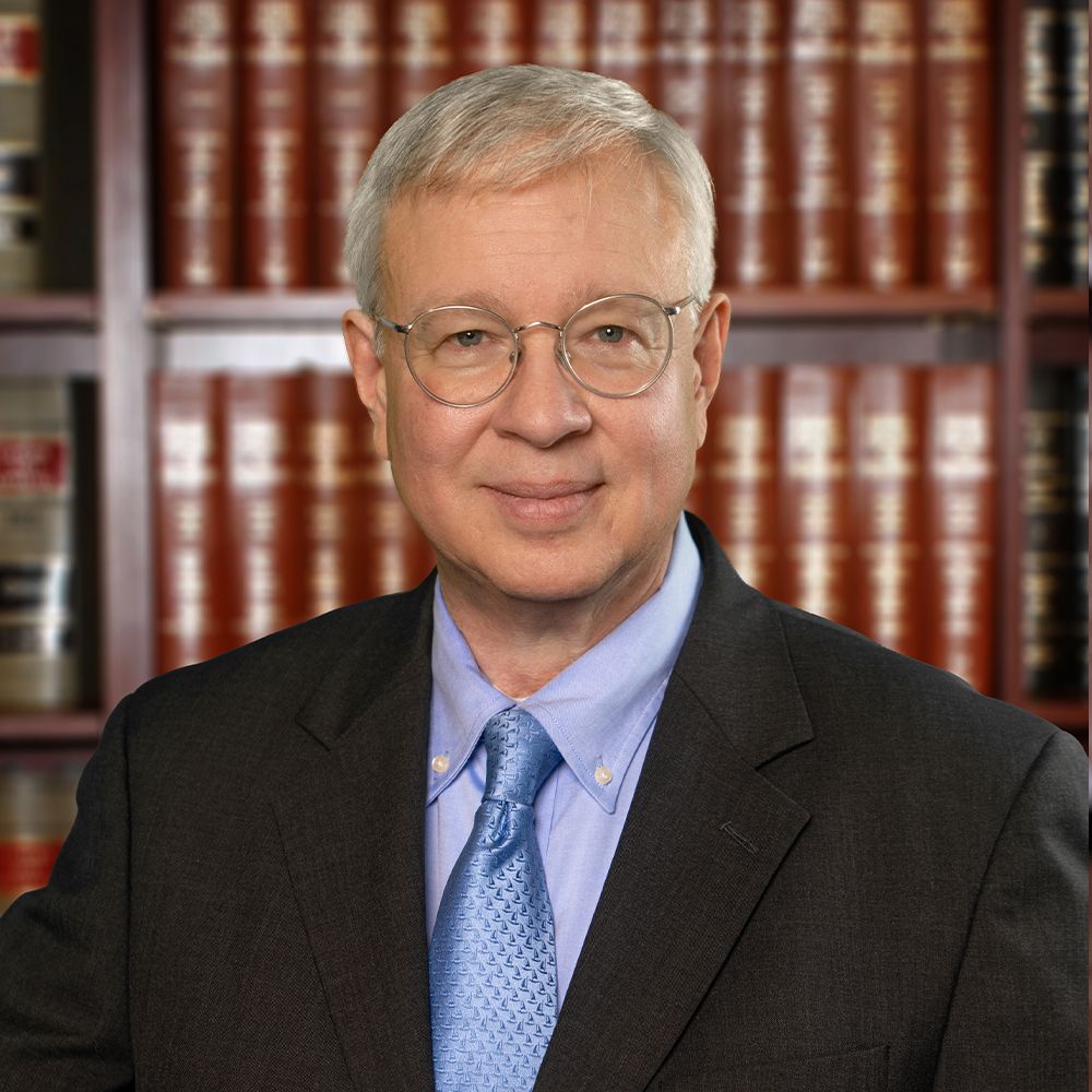 Elderly man in glasses and suit, standing in front of a bookshelf filled with law books.
