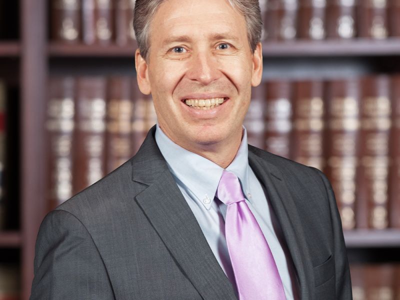 Smiling man in a suit with a pink tie, standing in front of a bookshelf filled with books.