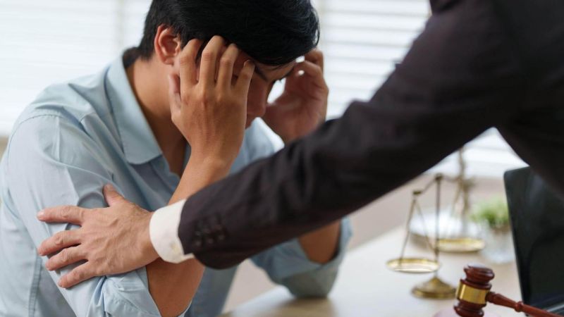 Man in a light blue shirt holds his head in distress while being comforted by a colleague in a room with legal objects (gavel and scales) on the table