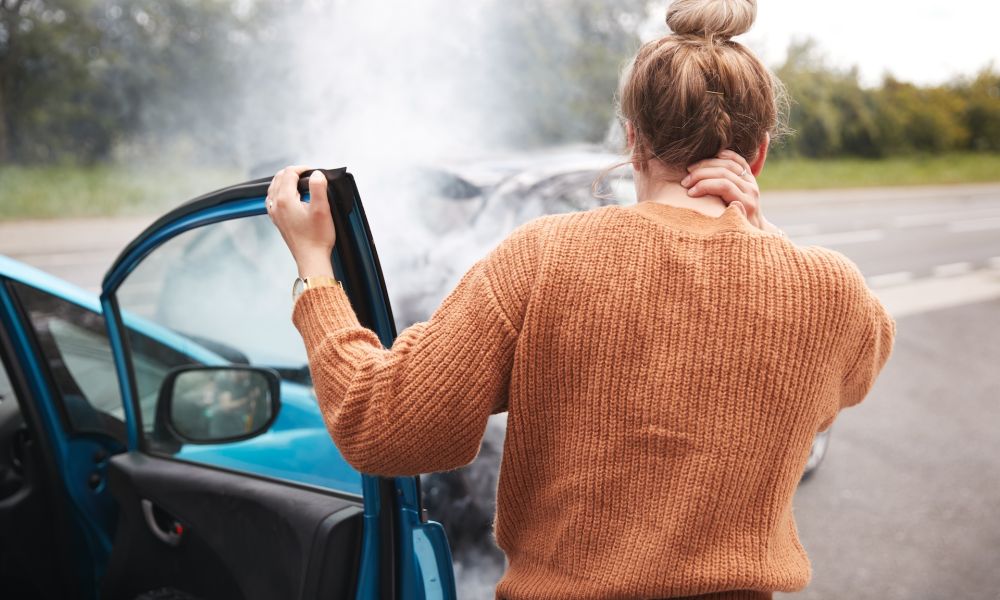 Woman with neck pain after car accident, standing beside blue vehicle on roadside.