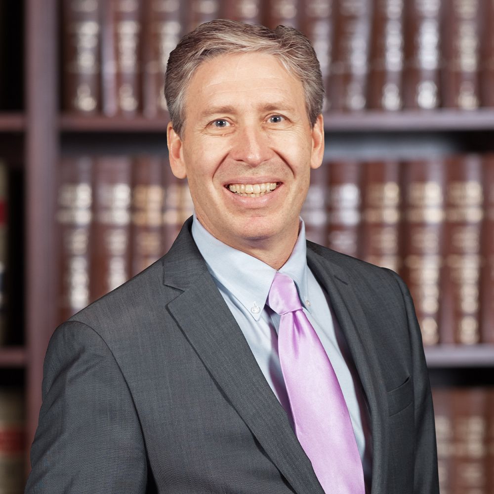Smiling man in a suit with a pink tie, standing in front of a bookshelf filled with books.
