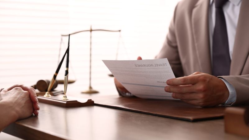 Two people in a formal setting exchange a document; a gavel and scales sit on the desk, suggesting a legal meeting.