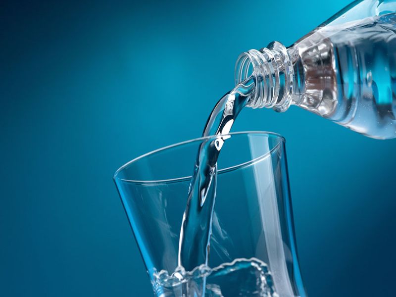 Pouring clear water from a bottle into a glass against a blue background, symbolizing hydration and purity.