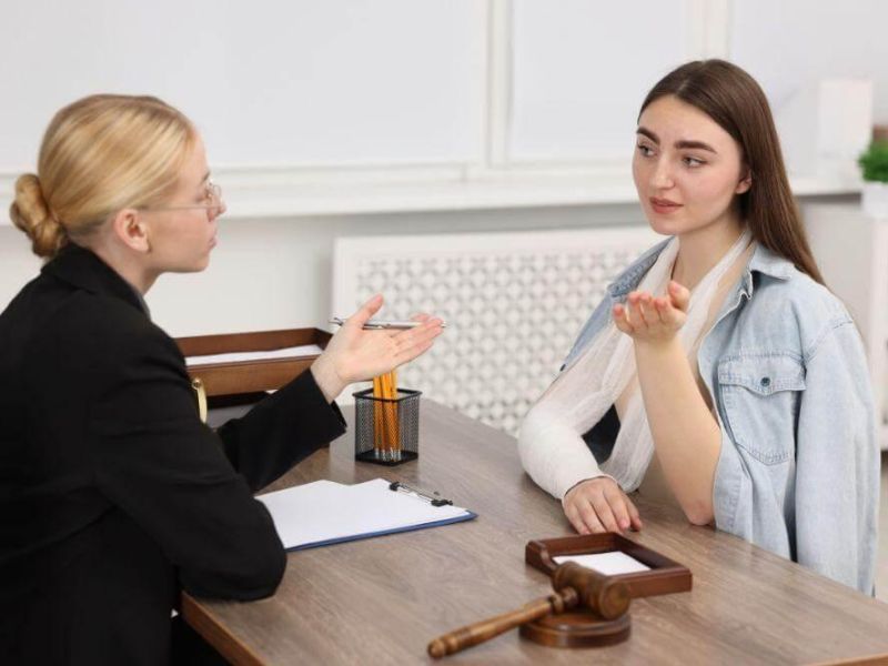Lawyer counseling a young woman with a cast on her arm at a desk, gavel and documents visible on the table, in a legal office.