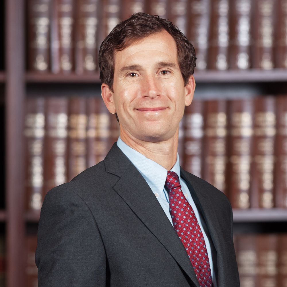 Man in a suit and red tie smiling in front of bookshelves filled with brown books.