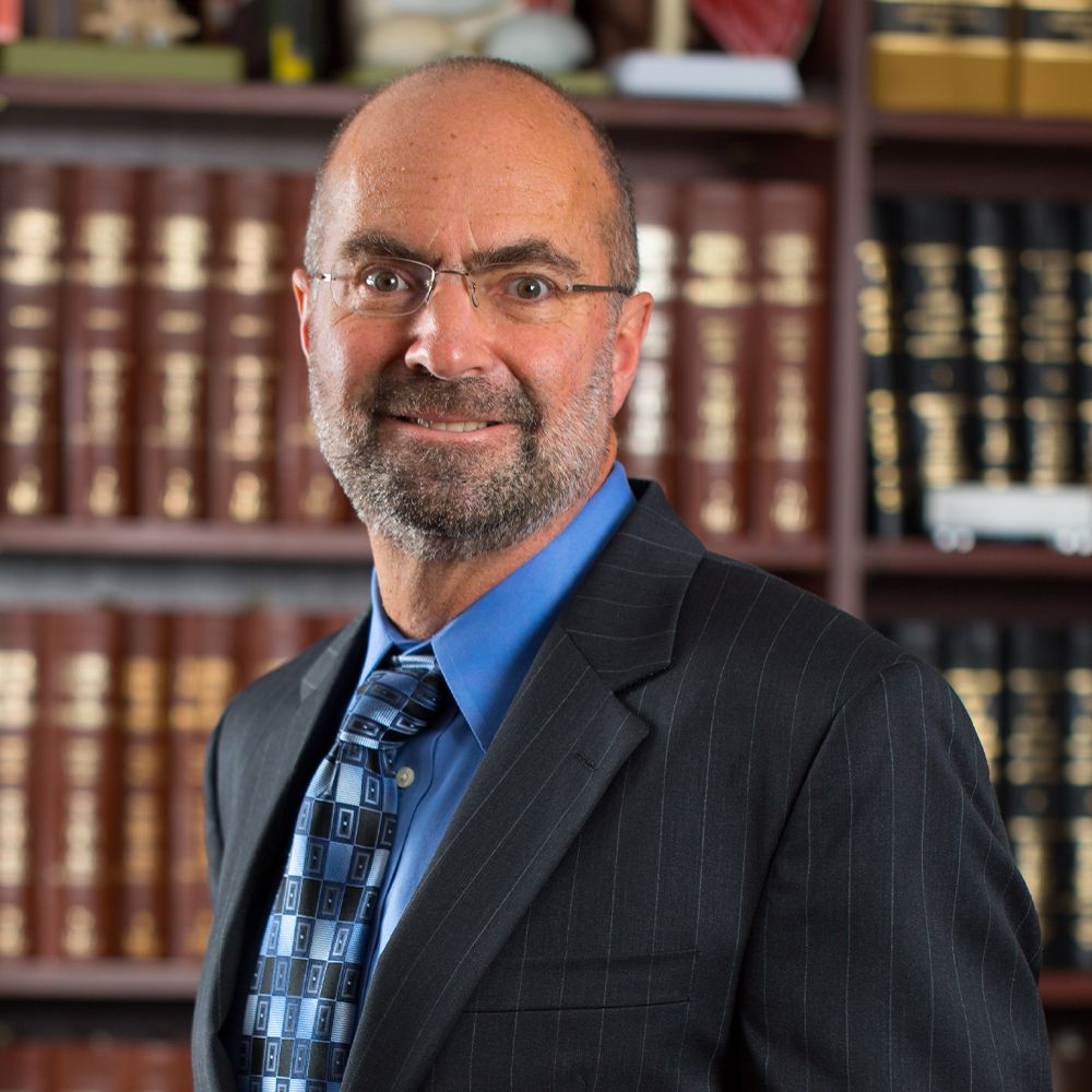 Professional man in suit with tie and glasses standing before bookshelves in office setting.