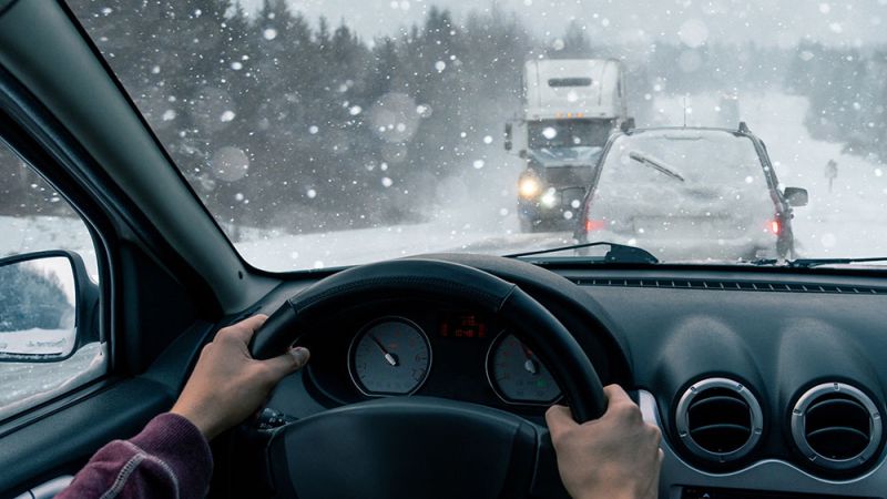 Driving in snowy weather, view from inside a car, surrounded by snow-covered vehicles and falling snowflakes.