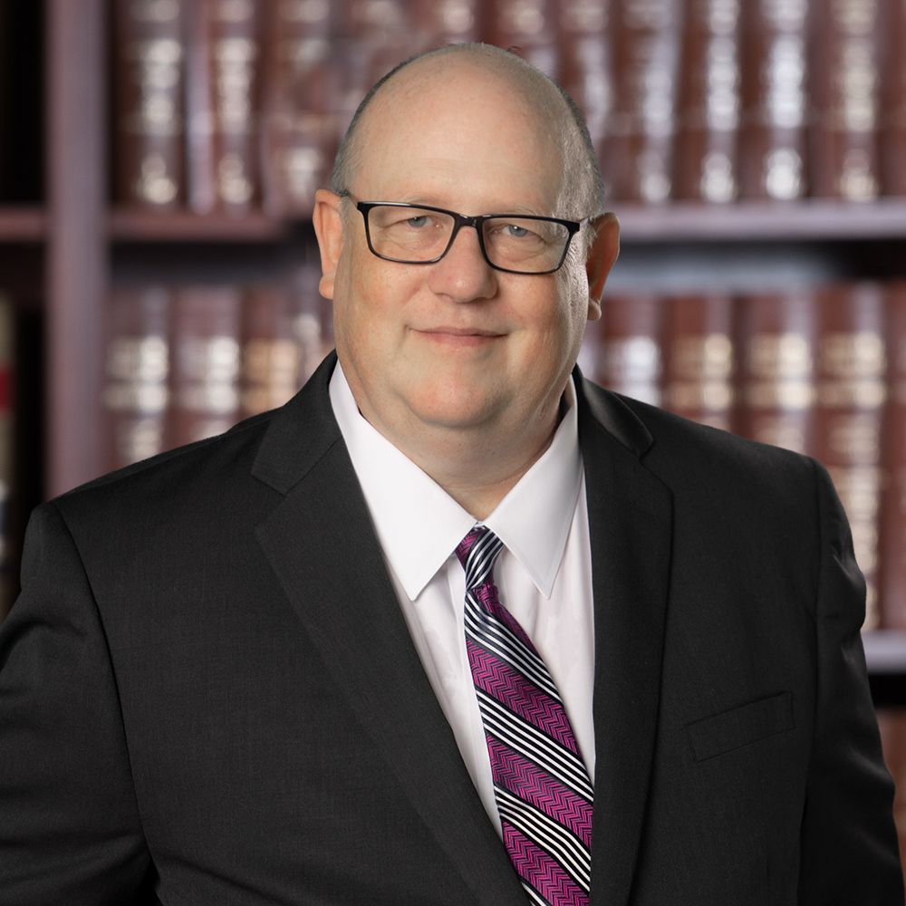 Man in a suit with glasses and a purple tie, standing in front of a bookshelf filled with books.
