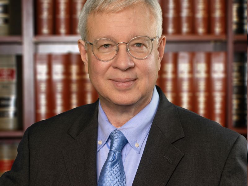 Elderly man in glasses and suit, standing in front of a bookshelf filled with law books.