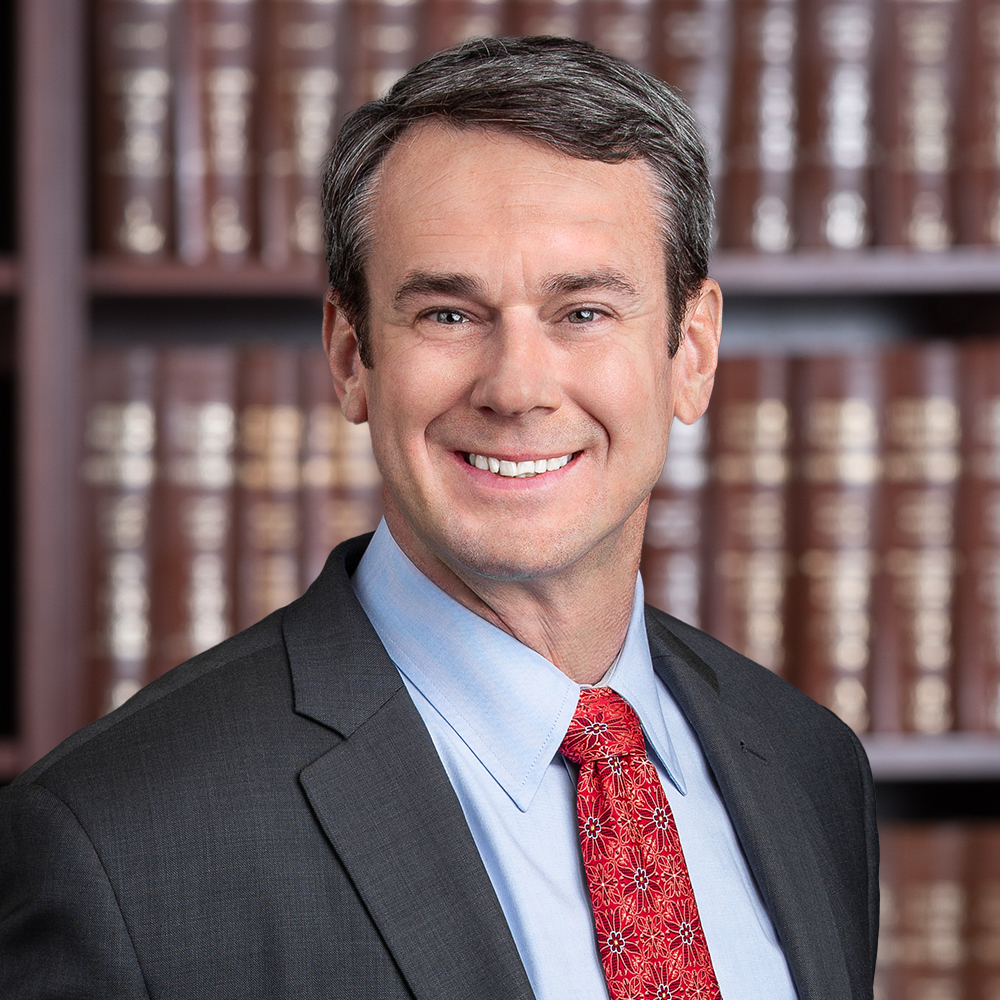 smiling man in a suit and red tie in front of a bookshelf filled with brown books