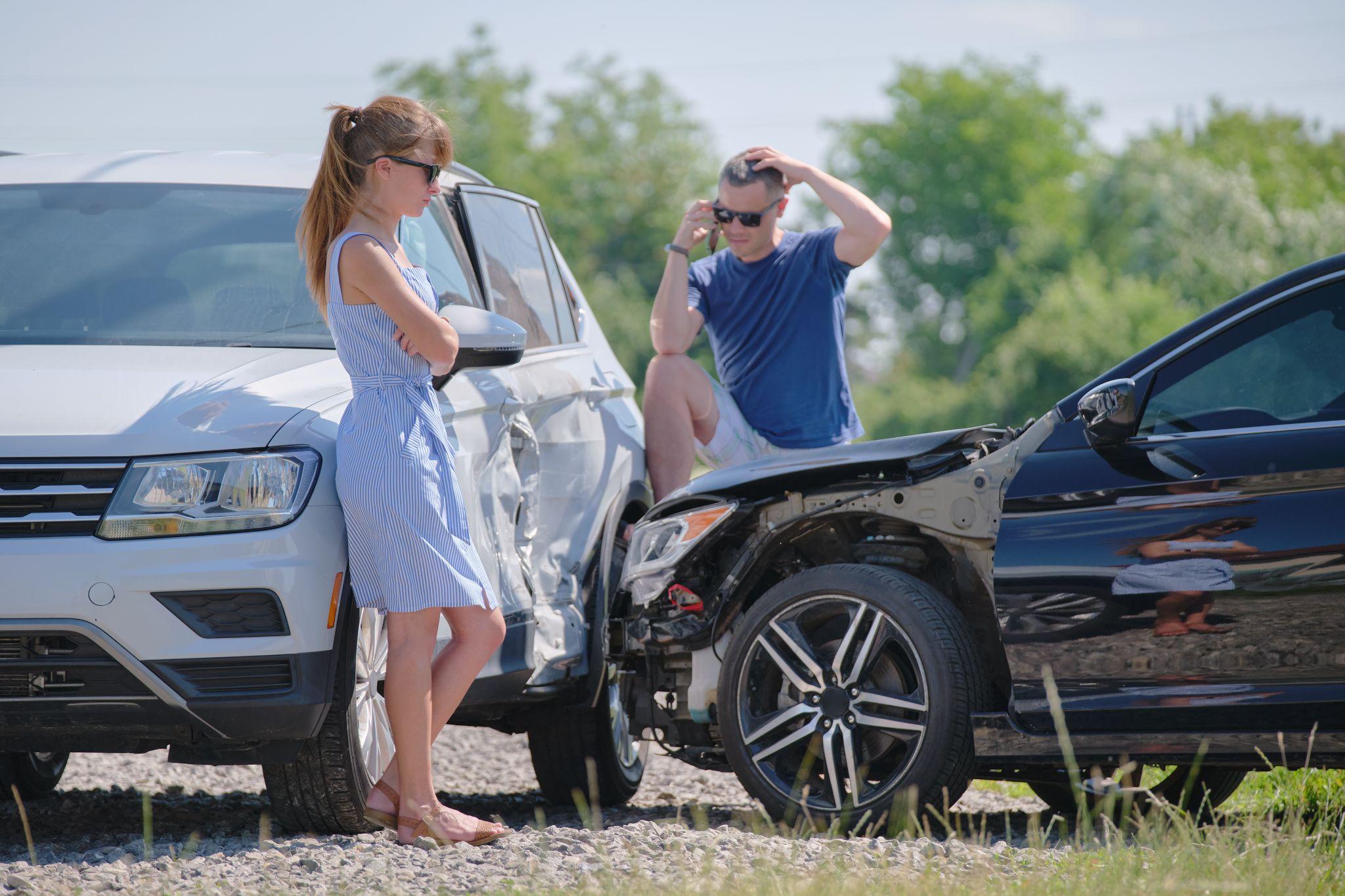 Two people standing near damaged cars after an accident, with a woman crossing her arms and a man making a phone call, both appearing concerned.