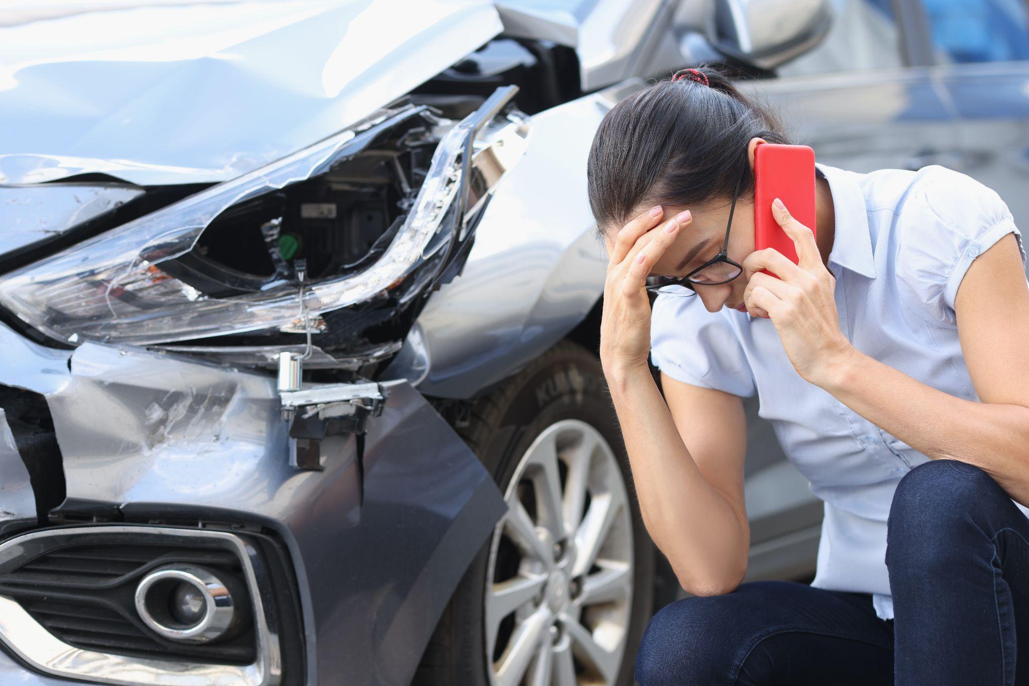 Upset woman making a phone call while sitting next to a badly damaged car after an accident