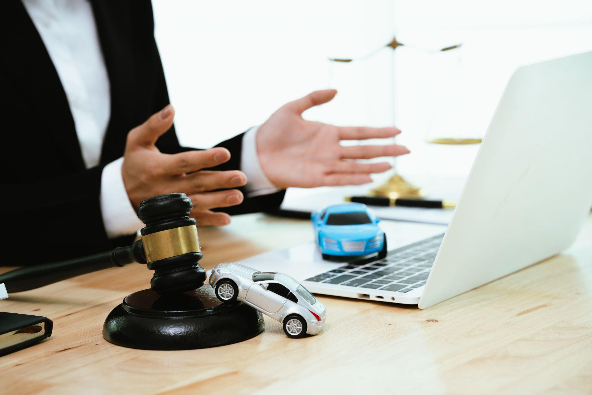 A lawyer gesturing with hands near a laptop, with a gavel and toy cars on the desk, symbolizing car accident or auto law cases.