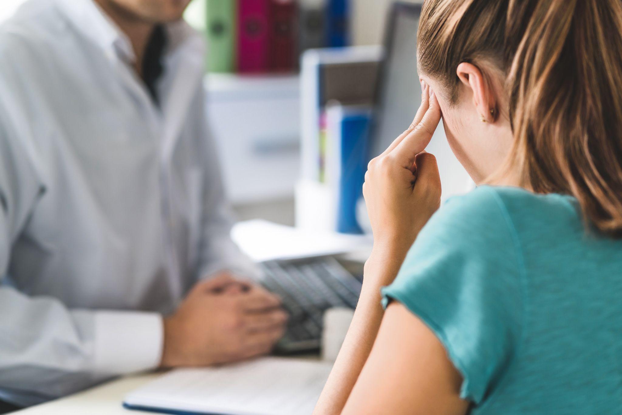 Woman at desk with doctor in background; she is touching her head, indicating a headache