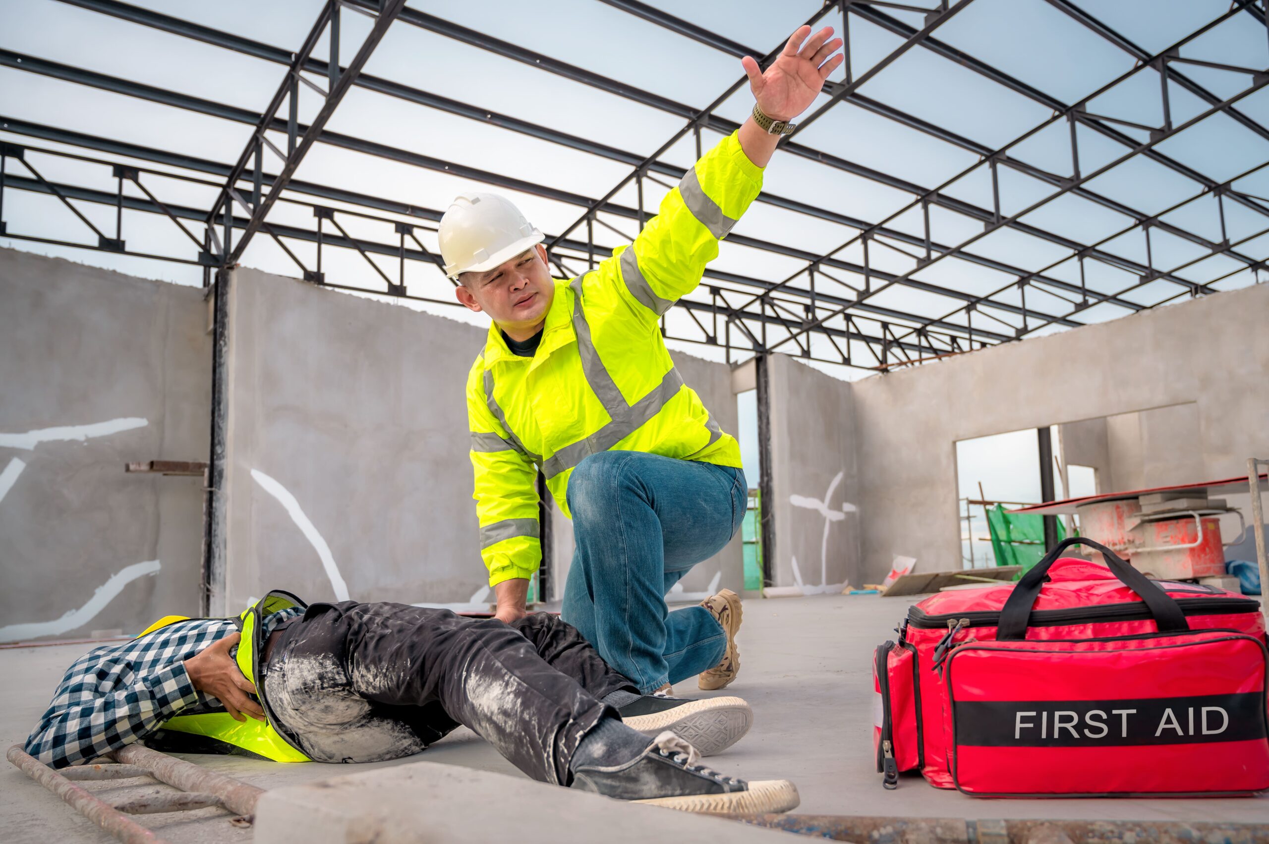 Construction worker in a neon safety jacket and white helmet tending to an injured person on a construction site, with a first aid bag nearby.
